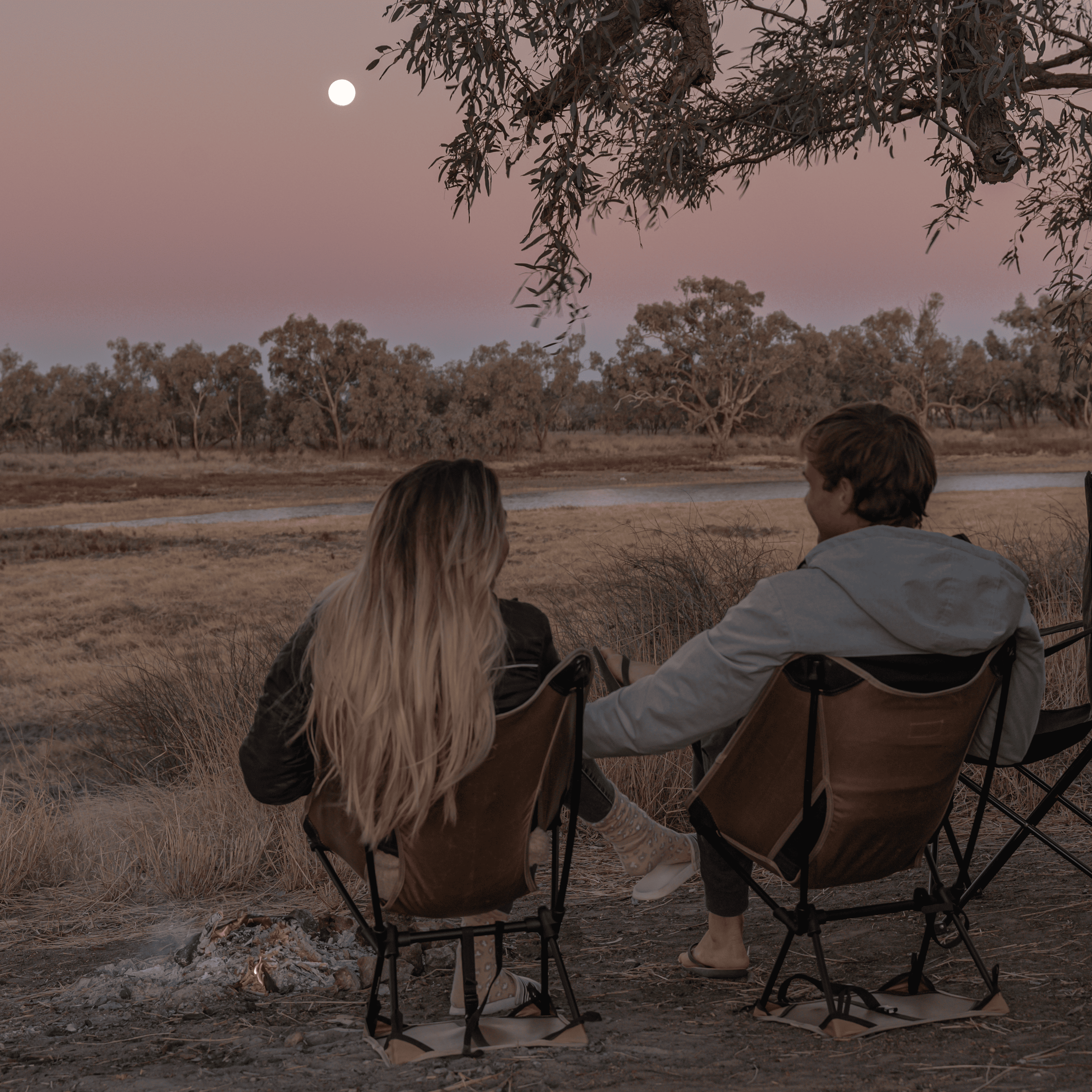 Wade and Dani sitting in their camping chairs in front of a firepit with the moon in the background. 