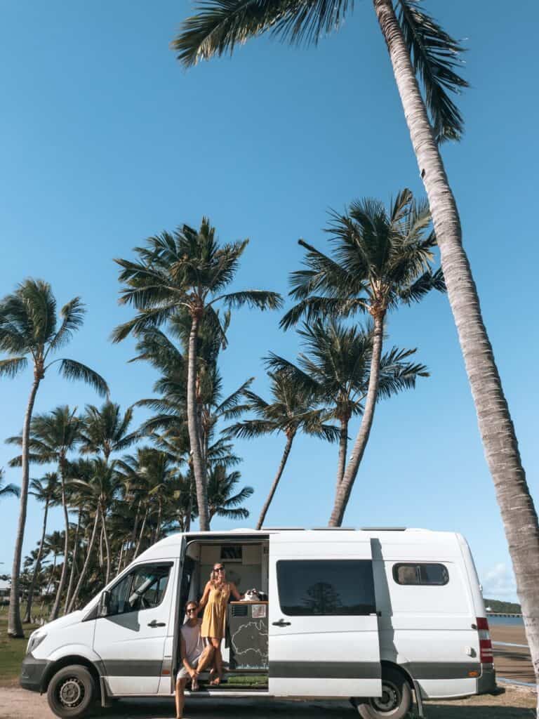 A campervan parked under some palm trees and Wade, Dani and their cat, Jon Snow, sitting on the door