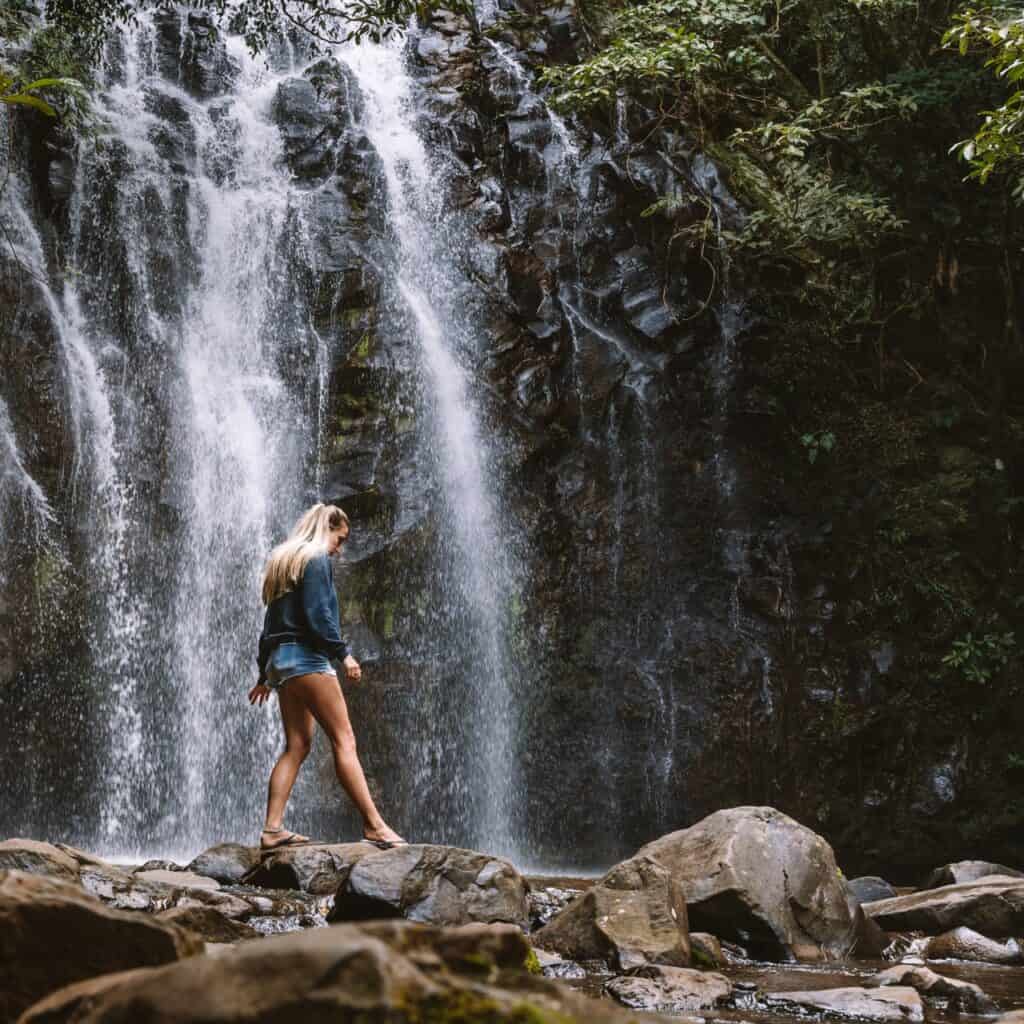 Dani crossing a small river by stepping on the stones at the bottom, in front of a waterfall