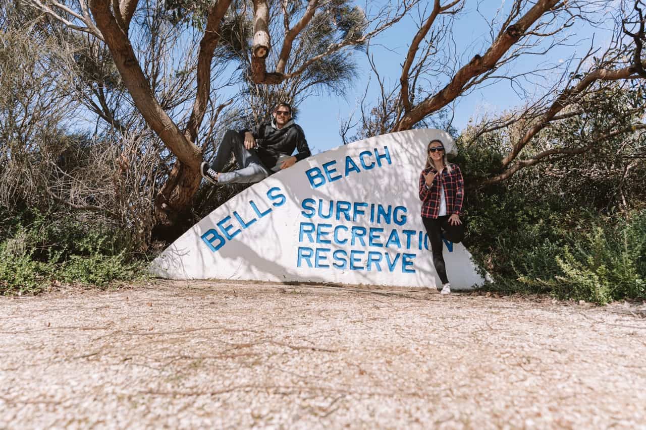 Dani and Wade standing in front of a sign that reads "Bells Beach - Surfing Recreation Reserve"