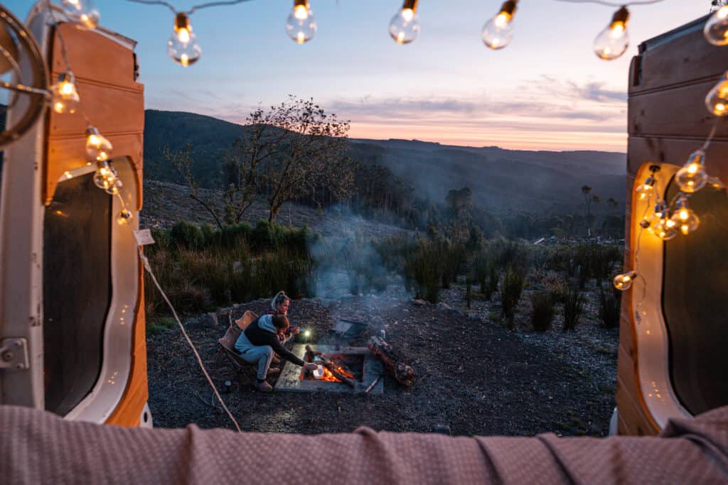 A view from the back of our campervan, with fairy lights on showing Dani and Wade by a firepit and a beautiful sunset