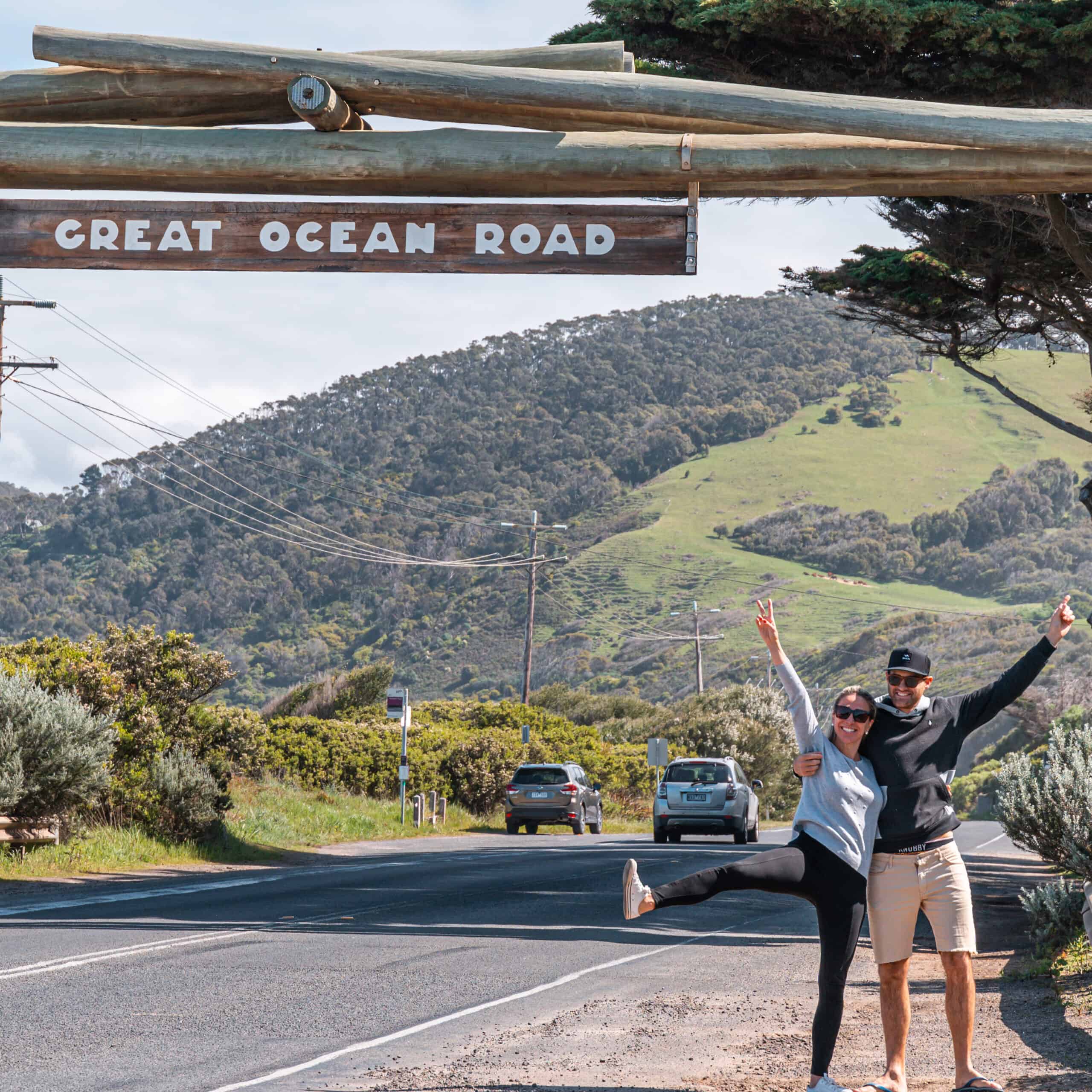 Wade and Dani standing under the Memorial Arch sign that reads Great Ocean Road. Dani has one foot on the floor and one up high, Wade and dani have their arms up