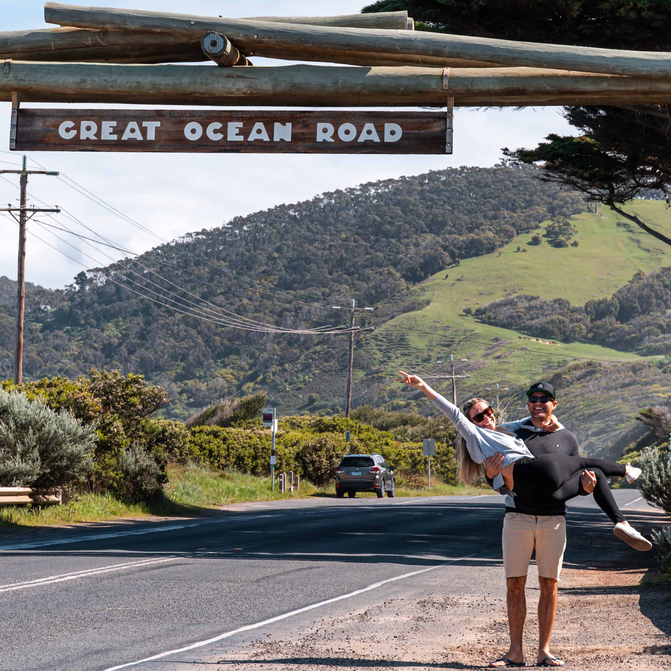 Wade and Dani standing under the Memorial Arch sign that reads Great Ocean Road. Wade has Dani in his lap like a baby