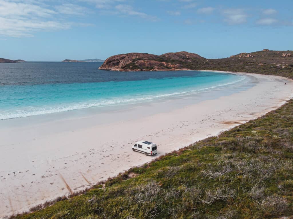 a van parked on the beach in Lucky Bay. The beach has a clean, white sand and turquoise water sheltered by rocks. 