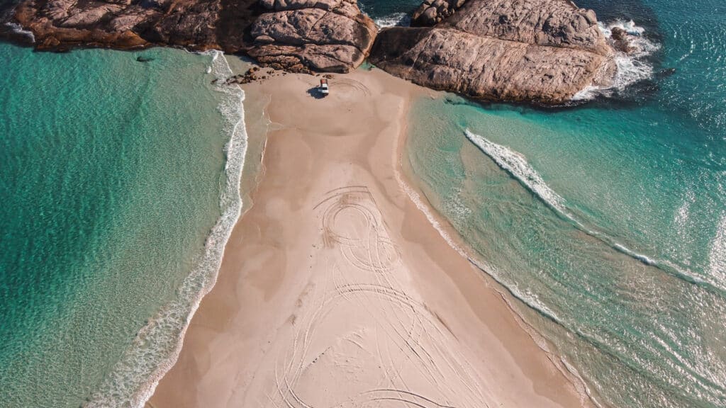 A drone picture at Wylie Bay Beach. You see a strip of sand in the middle with turquoise water on both sides and a car parked next to the rocks 