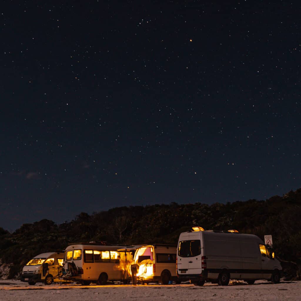 A few vans parked on the beach in Wharton Beach at night. The sky is full of stars